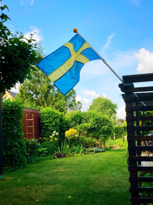 Svenska flaggan vajar på en stång i en grönskande trädgård med blommor och träd.