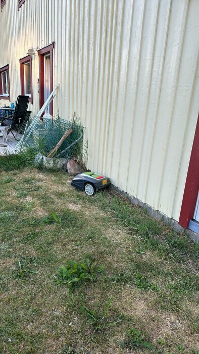 Robotgräsklippare nära en gul husvägg med synlig grävning för plattläggning och avklippt kabel.