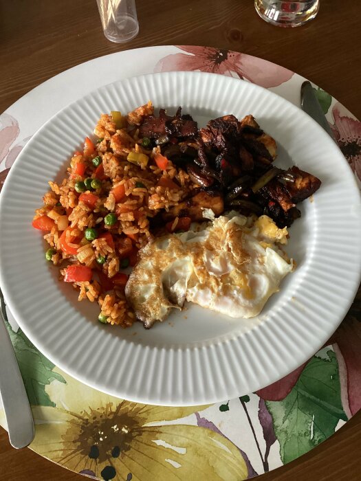 Tallrik med stekt ris, grönsaker, kyckling och stekt ägg, serverat på ett mönstrat bord.