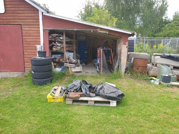 Garagefasad med ny panel på högra sidan och dörrar målade i faluröd svart, omgivet av diverse föremål.