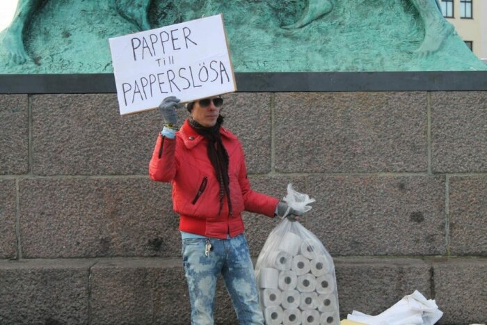 Person i röd jacka håller skylt med text "Papper till papperslösa" och en säck med toalettrullar.