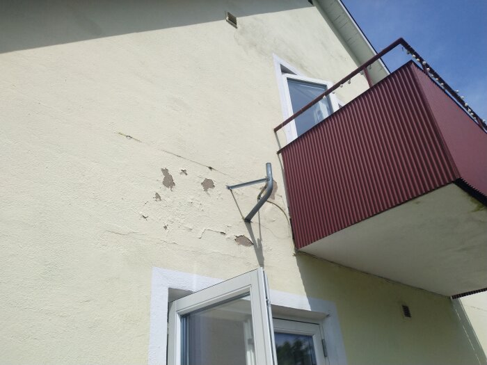 Delar av skalad färg på husvägg under balkong, tom parabolhållare på vägg.