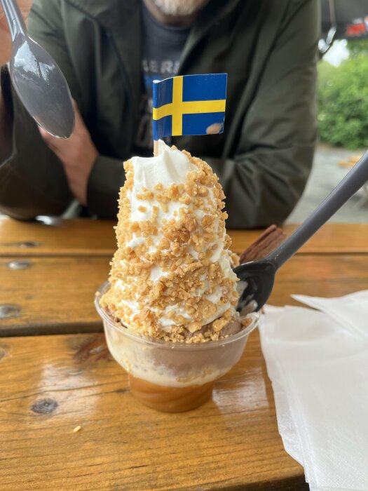 Glass med vispgrädde och karamelliserade nötter toppad med en svensk flagga, serverad i en plastbägare.