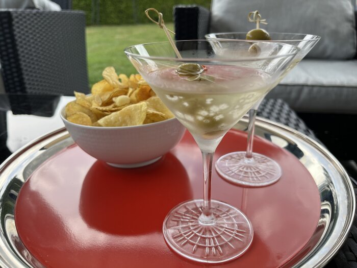 Martini med oliv och skål med chips på ett rött brickbord utomhus.