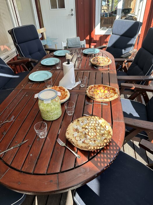 Fyra olika pizzor på ett träbord utomhus, dukat med tallrikar och glas.