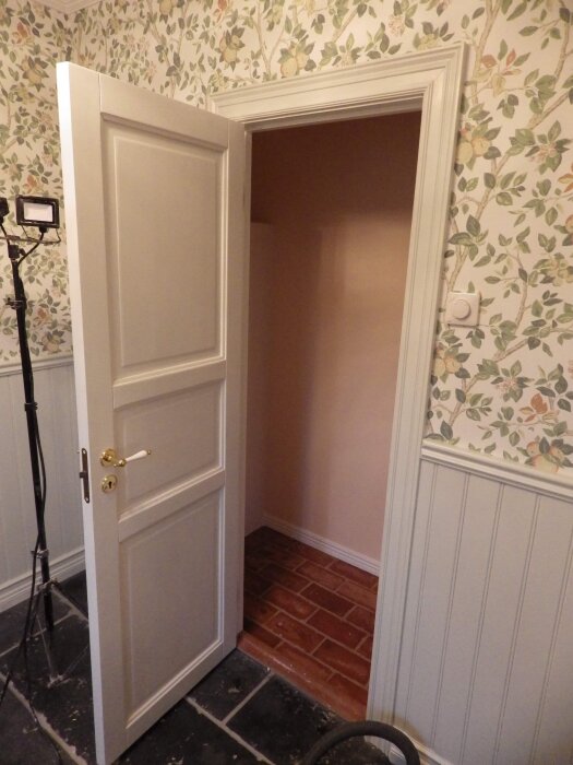 Öppen vit dörr som visar övergången mellan en rum med tapet och pärlspont till ett annat med brunmålad vägg.