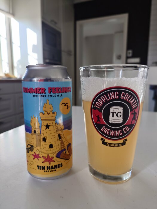 Ölburk med texten "SUMMER FEELINGS DDH HAZY PALE ALE" och glas med ljust öl på köksbänk.