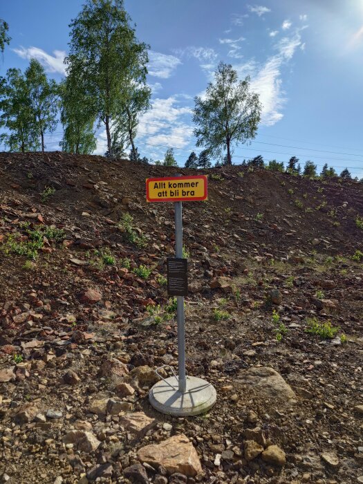 Skylt med texten "Allt kommer att bli bra" framför en backe med stenar och träd under en blå himmel.