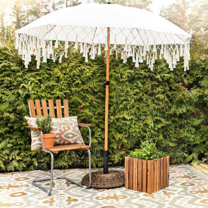 Sekelskiftesinspirerad uteplats med vit parasoll, trästol, krukväxter och mönstrad matta.