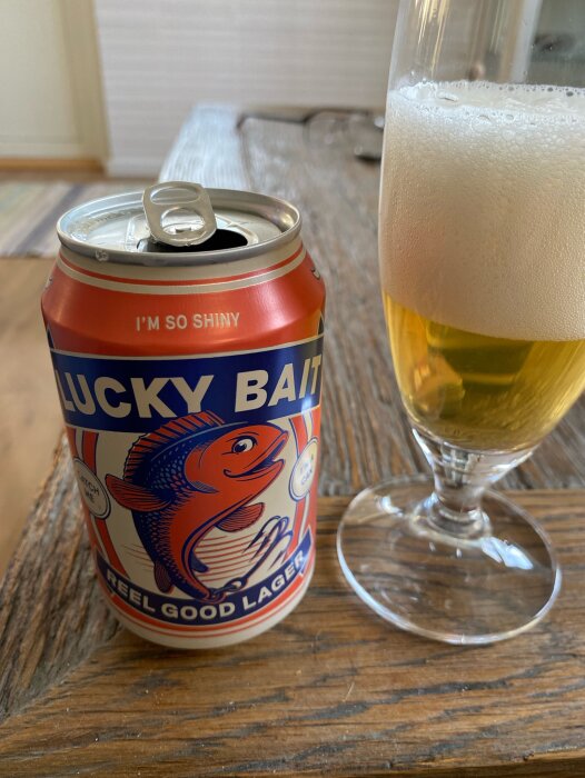 Öppnad ölburk med texten "LUCKY BAIT REEL GOOD LAGER" och ölglas med skum på träbord.