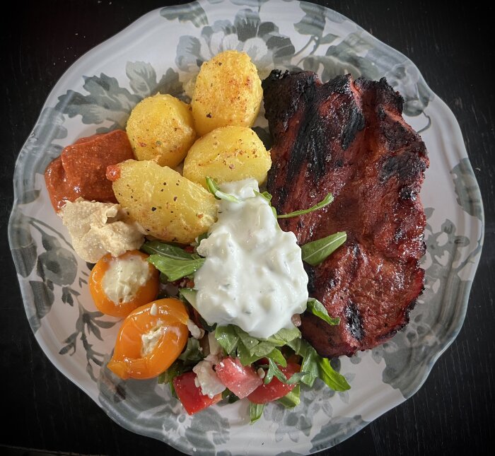 Grillad fläskkarré med kryddad potatis, sallad, hummus, tomat/chiliröra och tsatsiki på en tallrik.