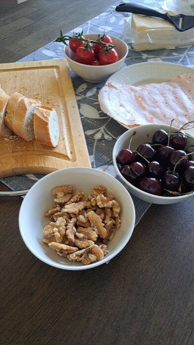 Skärbräda med skivat bröd, skål med körsbär, valnötter, tomater och ost på köksbord.
