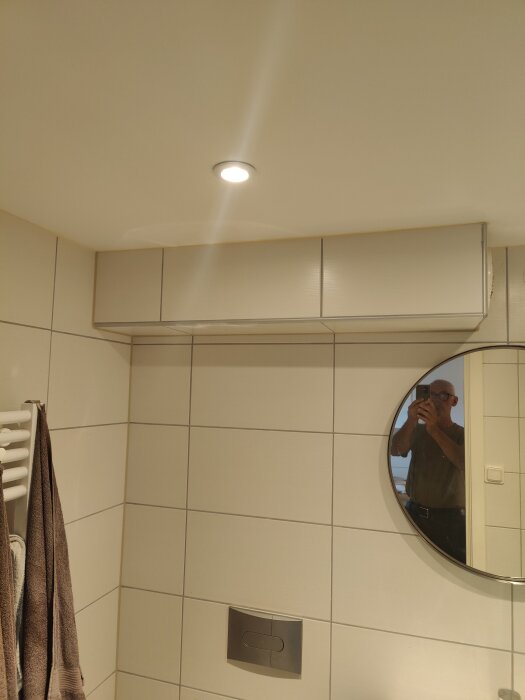 Badrumsinteriör med vita kakelväggar, spegel och taklampa, reflektion av person som tar fotot.