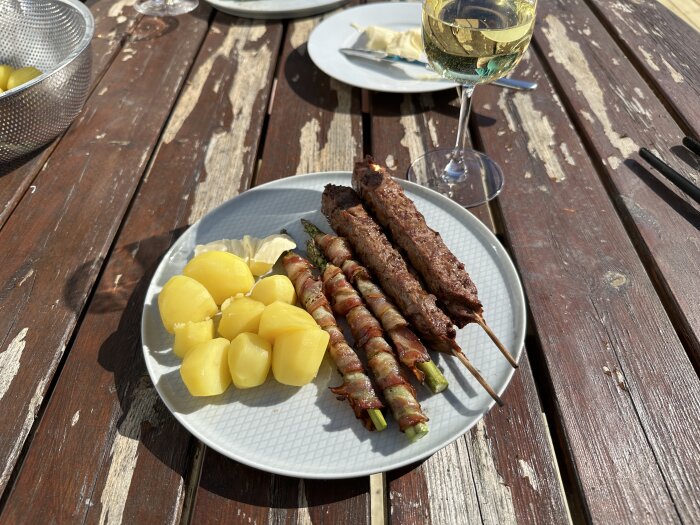 Måltid på trätallrik med kebabspett, baconlindade sparris och kokt potatis på en slitna träaltan.