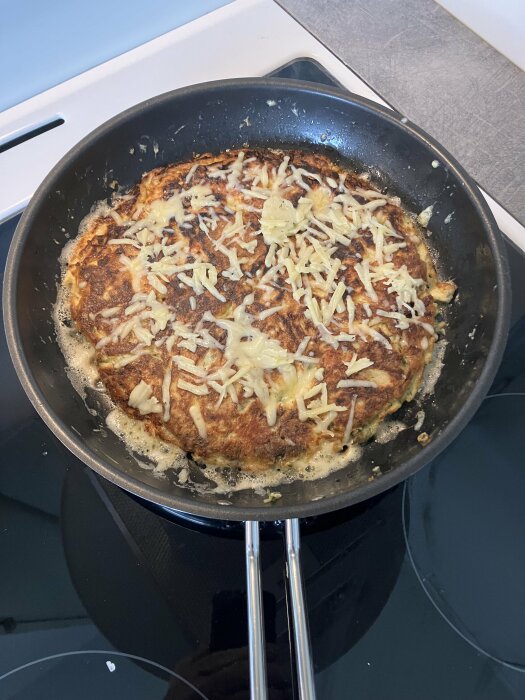 Frittata med lax och potatis i en stekpanna på spishäll, överströdd med riven ost.