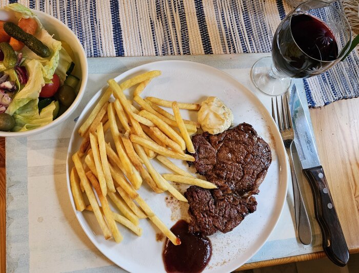 Middagstillbehör bestående av en tallrik med grillat kött, pommes frites, bearnaisesås, sallad och ett glas rödvin.