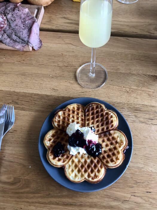 Våffla med grädde och sylt på en tallrik, skinka på skärbräda och ett glas med ljusgul dryck vid en träbordsskiva.