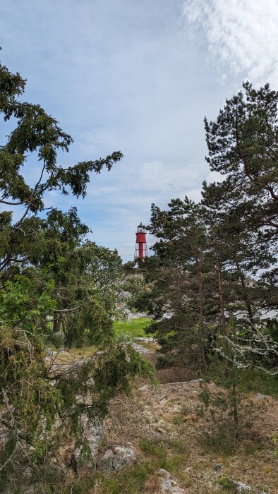 Röd och vit fyr synlig mellan gröna träd och över klippigt landskap mot en blå himmel med moln.