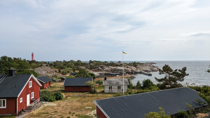 Röda trähus, klippor, svensk flagga och en fyr vid havet under en molnig himmel.