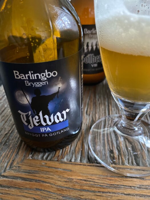 Tre ölflaskor på ett träbord, förgrunden visar Barlingbo Tjelvar IPA med synlig text, bakgrunden visar andra öl och ett glas med skum.