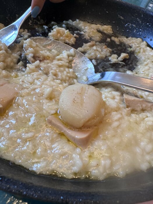 En portion risotto med en pilgrimsmussla och anklever ovanpå, redo att serveras, tillagad i en stekpanna.