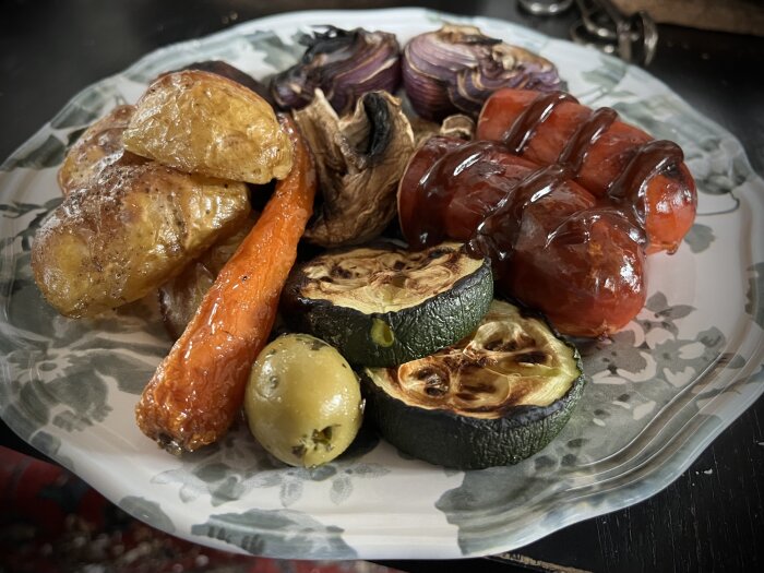 Grillspett med bratwurst och grillade grönsaker som squash och morötter på en mönstrad tallrik.