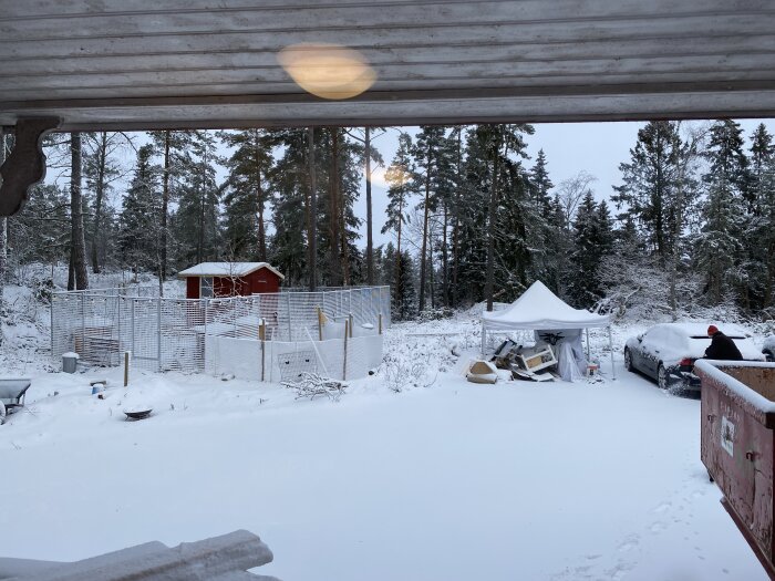 Vinterlandskap av en gårdsplan med en gammal hönsgård omgiven av stängsel och snö, samt diverse byggmaterial och fordon i förgrunden.