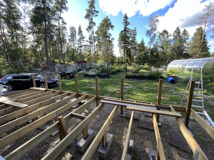 Altan under konstruktion med träreglar och stolpar, omgiven av trädgård och fordon.