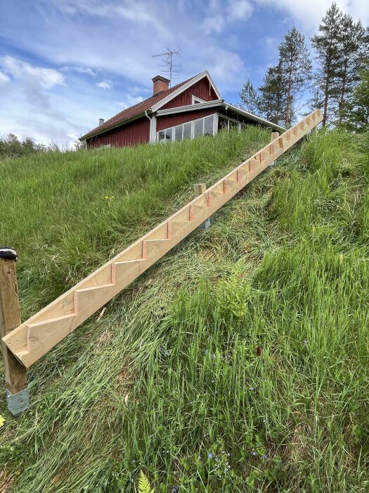 Nybyggd trätrappa på sluttande gräsmark med ett rött hus i bakgrunden.