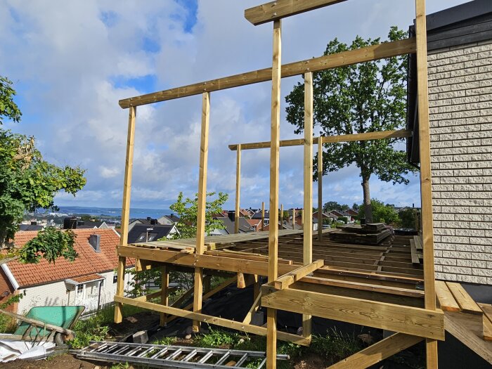 Träkonstruktion av ett pågående altanbygge med synliga stolpar och deras fästpunkter mot bakgrund av hus och himmel.