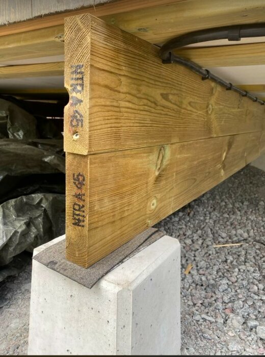 Stödbjälke i trä placerad ovanpå en betongpelare vid en husgrund, möjlig säkerhetsrisk.