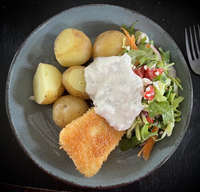 Panerad torsk med dillsås, kokt färskpotatis och blandad grönsallad på en grå tallrik.