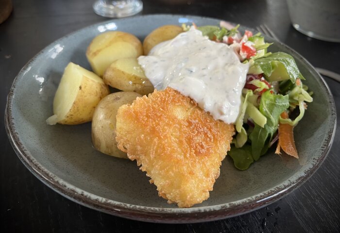 Panerad torsk med dillsås, kokt färskpotatis och sallad på en keramiktallrik.