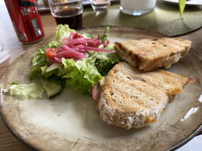 Grillad smörgås halverad och serverad med sallad och inlagd rödlök på en keramiktallrik med en burk Coca-Cola i bakgrunden.