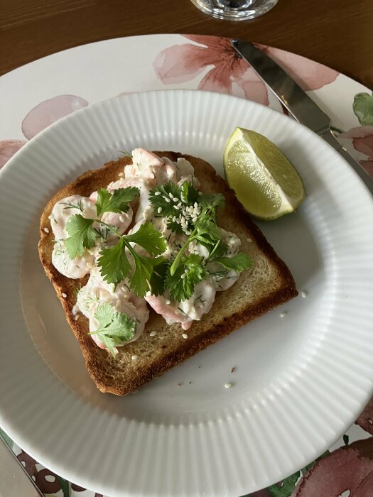 Skagenröra på rostat bröd toppad med koriander och sesamfrön, serverad med limeklyfta på blommig tallrik.