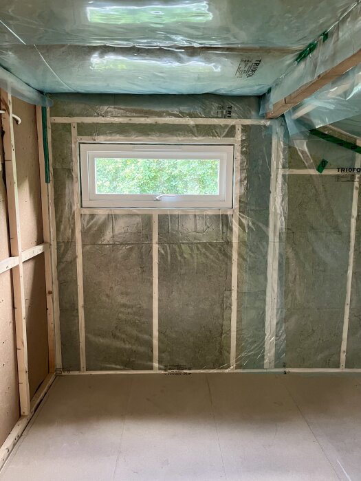 Utrustat sovrum under renovering med nytt litet fönster i ögonhöjd, vindpapp och isolering syns.