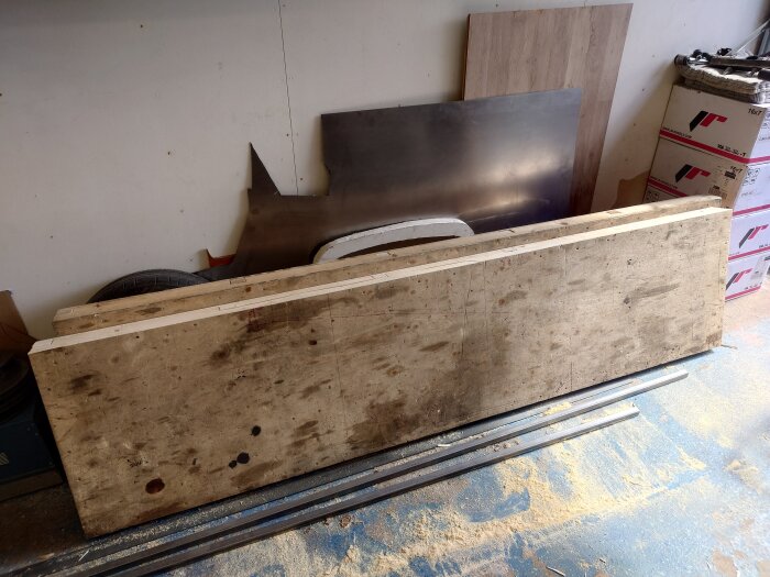 Lång, använd bänkyta lutad mot en vägg i ett garage omgiven av byggmaterial och verktyg.