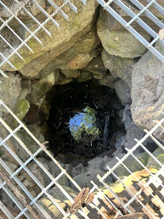 Gammal grävd vattenbrunn sedd genom ett rostigt gallertak, med vatten och stenar synliga nertill.
