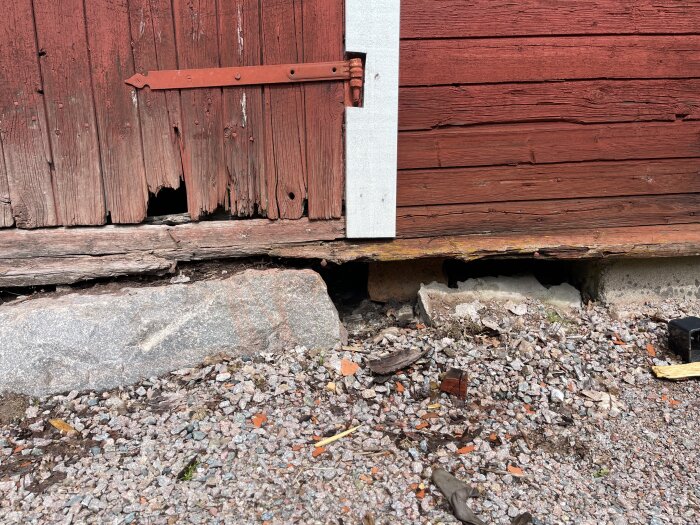 Skadad träpanel och röta vid nedre delen av en rödmålad ladas hörn med grus och stenar framför.