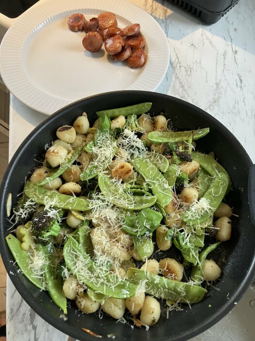 Stekt gnocchi med broccoli och sockerärtor, toppat med riven ost, och skivor av kolbacs på en tallrik bredvid.