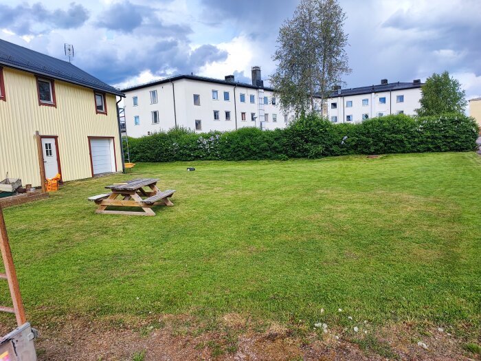 En gräsmatta med en picknickbänk i förgrunden, omgärdad av en gul byggnad med förråd och vita bostadshus i bakgrunden under en molnig himmel.