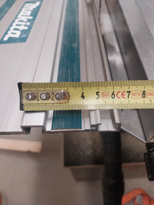 Mätning av aluminiumprofil med måttband visar en längd på cirka 5,6 cm, profilerad aluminiumport med logotypen "Makita" synlig i bakgrunden.