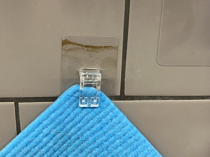 Transparent plastkrok monterad på en kaklad vägg, som håller en blå rengöringsduk.