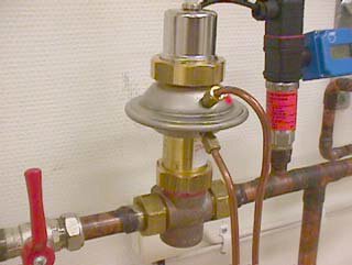 En tryckregulatorventil installerad på en kopparrörledning i ett VVS-system, omgiven av olika anslutningar och ventiler.