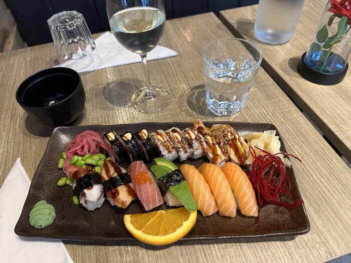 En tallrik med sushi, inklusive lax, tonfisk, avokado, wasabi, inlagd ingefära och edamame. Ett glas vitt vin och vatten på bordet.