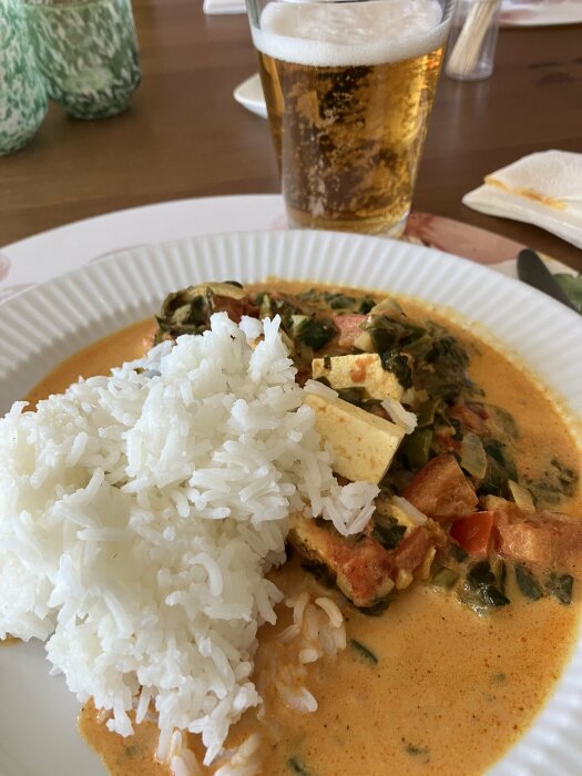 En tallrik med ris och grön currygryta samt ett glas öl i bakgrunden på ett träbord.