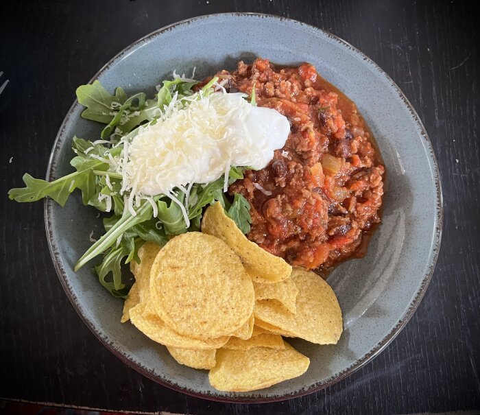 En tallrik med chili con carne, ruccola, nachos, vitlökssås och strödd ost på topp.