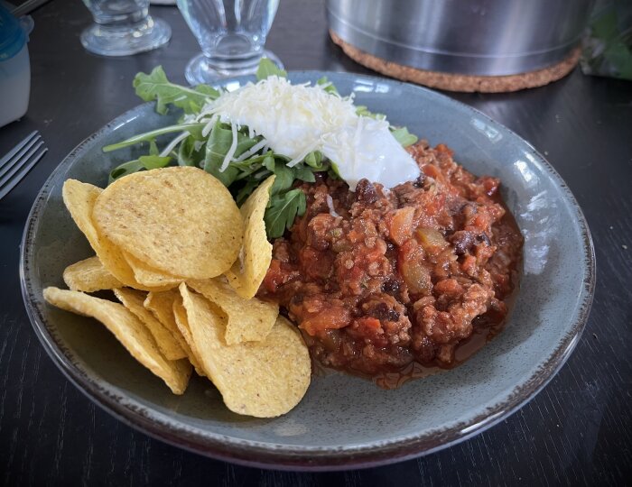 En tallrik med chili con carne, ruccola, vitlökssås, strödd ost och en hög med nachos på ett mörkt bord.