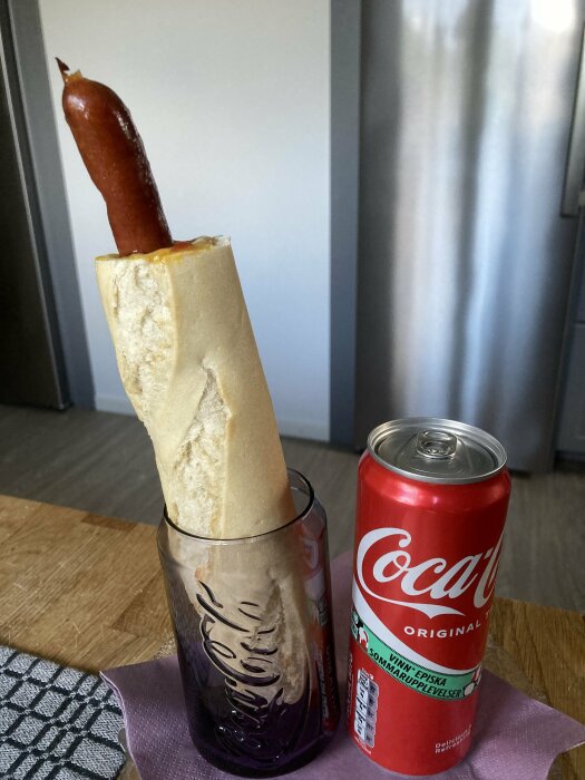 En baguette fylld med korv, ketchup och senap står i ett glas bredvid en burk Coca-Cola på ett träbord i köket.