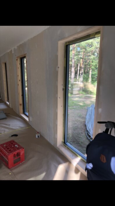 Fönster med ram av obehandlat trä i ett rum under renovering, skyddspapp på golvet och målarburk på fönsterbrädan, skog syns genom fönstren.